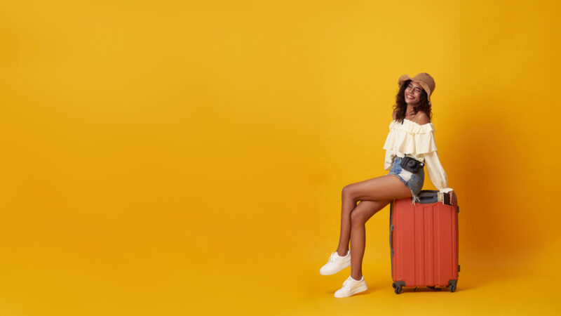 Young pretty african woman tourist smiling sitting on luggage with camera in hand isolated on yellow background.